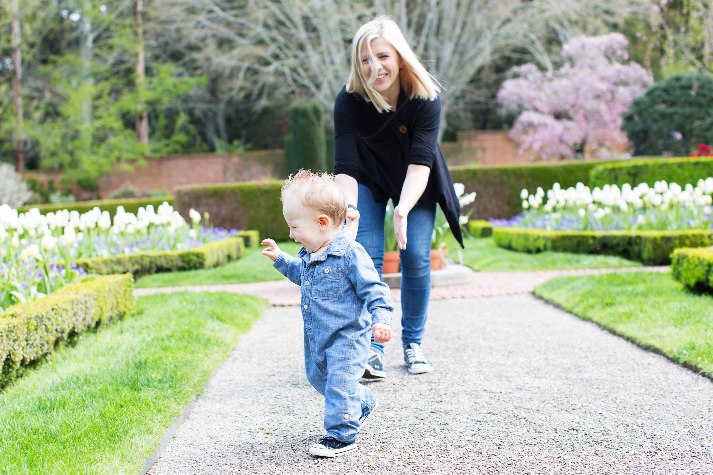 Laughing cute toddler boy running away from young mother in park with flowers