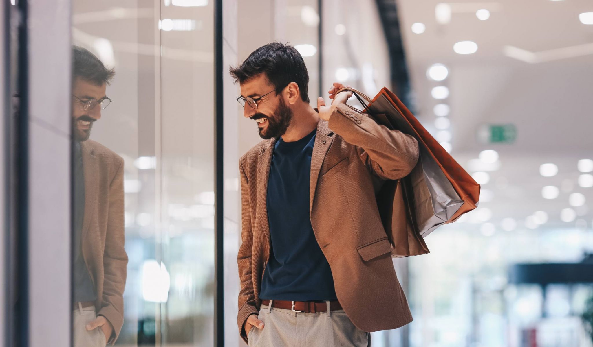 A man walks through a shopping mall with a smile on his face and bags in his hand and looks at the shop windows